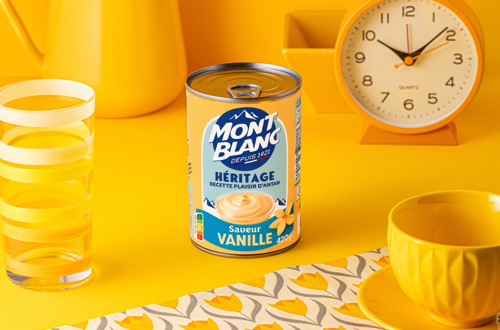 Boîte Mont Blanc Héritage saveur vanille sur une table jaune, avec verre, tasse et horloge rétro, au format paysage.